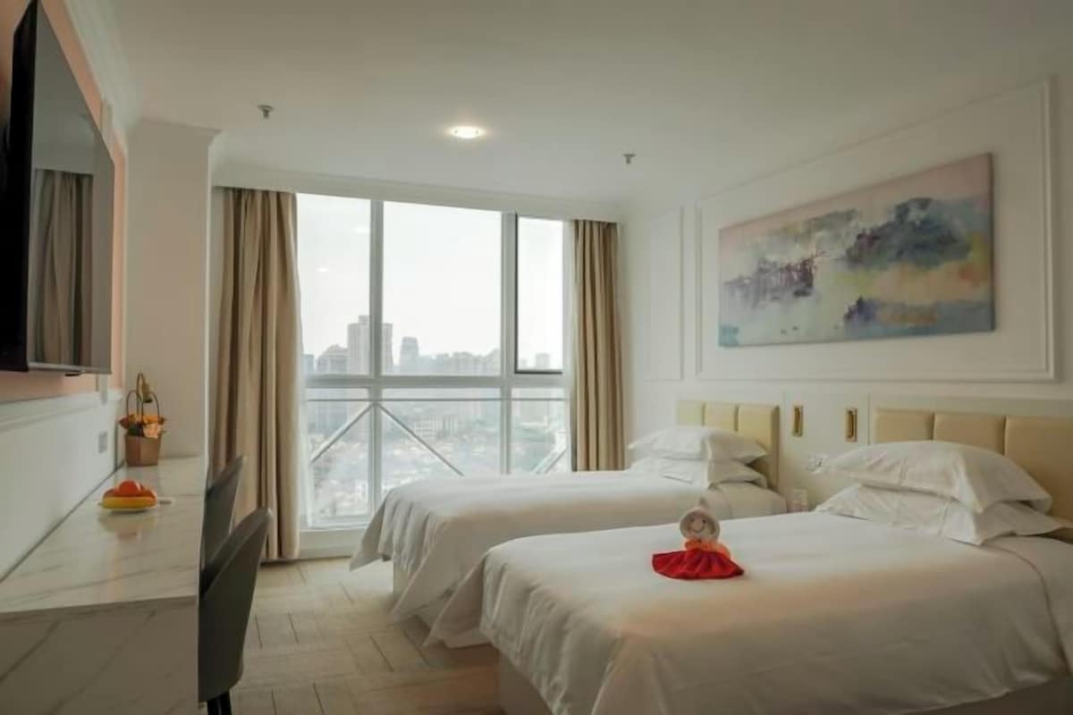 A hotel room with two beds, a large window with a city view, a painting on the wall, and a flat-screen TV on the left.