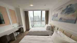 Habitación de hotel con dos camas, un escritorio y un amplio ventanal con vista a la ciudad.