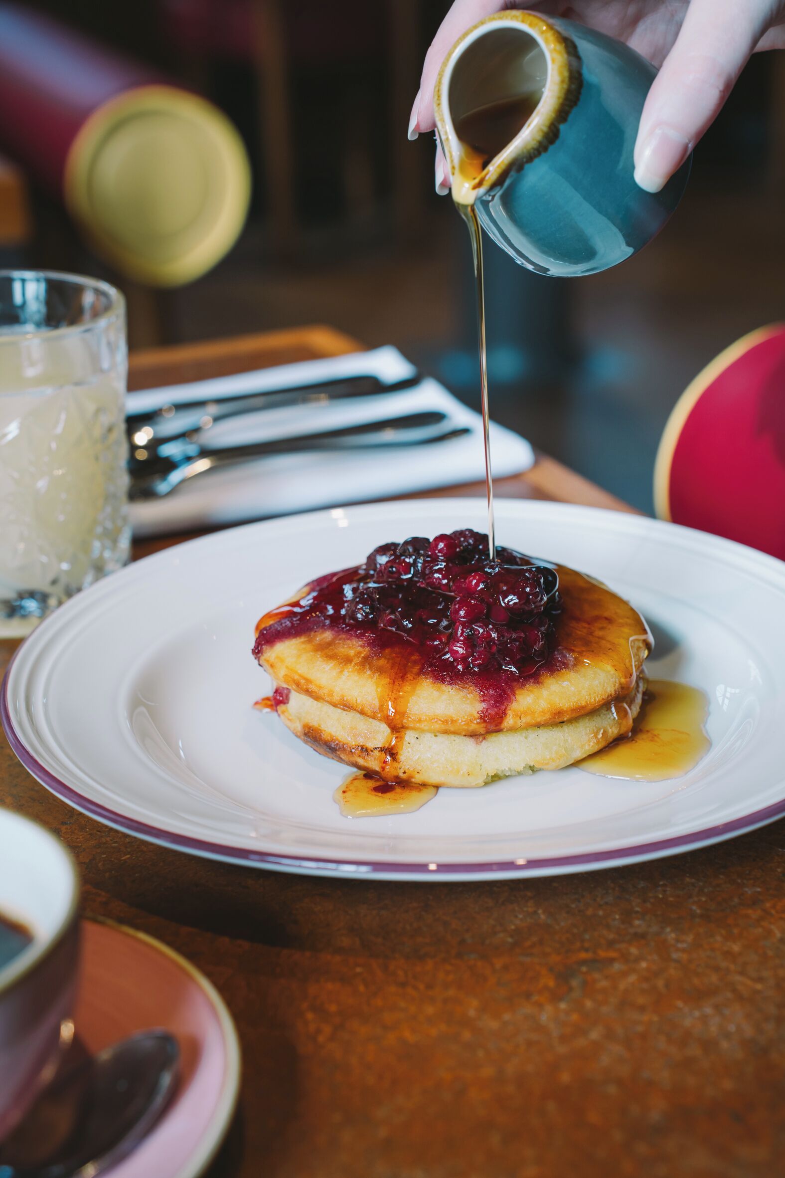 A plate with pancakes topped with berries and syrup, a glass of a clear liquid, and a spoon on a saucer.