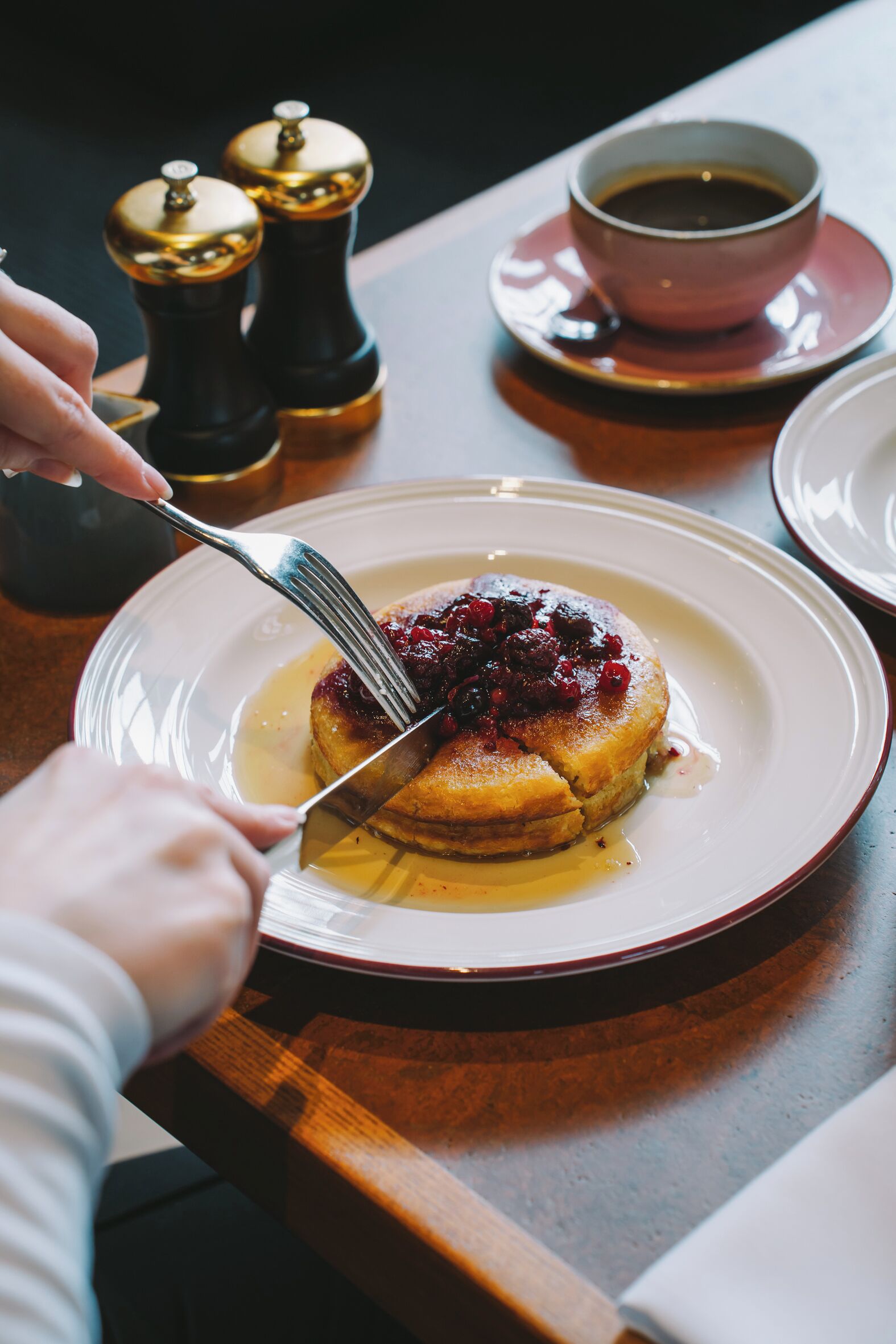 Une assiette de crêpes garnies de fruits rouges et nappées de sirop, une tasse de café, et des salières et poivrières posées sur une table.