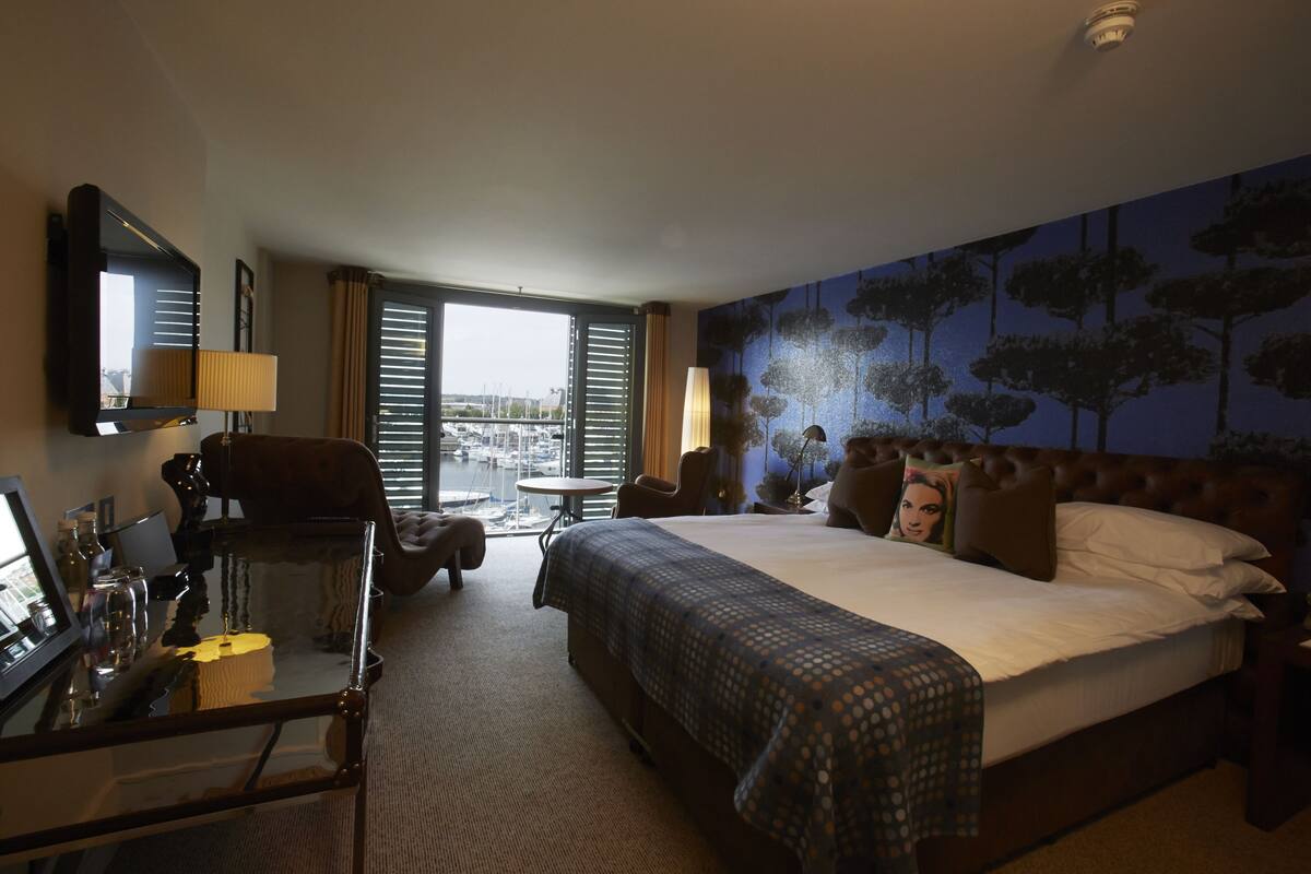 Ein Hotelzimmer mit einem großen Bett, einem Schreibtisch mit Lampe, einem Sessel und Blick auf die Stadt durch große Fenster.
