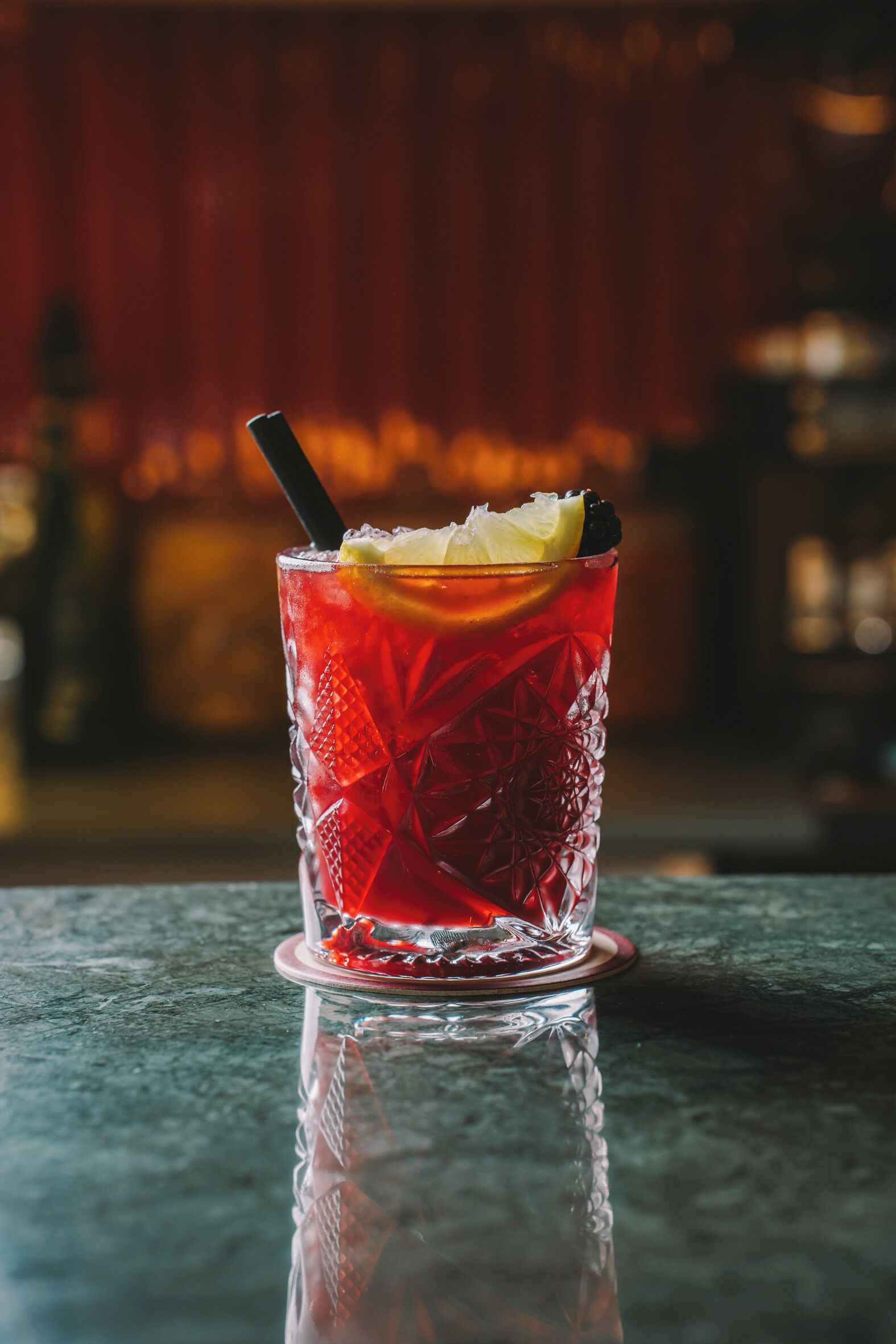 Un verre transparent contenant une boisson rouge, agrémenté d’une tranche de citron et d’une paille noire, posé sur une surface réfléchissante.