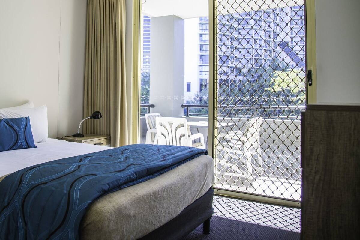 A hotel room with a bed, a nightstand, a desk, a chair, and a balcony with a view of buildings.