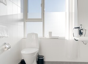 A modern bathroom with a toilet, a trash bin, a towel rack, a window with a view, and a hairdryer on the sink. - Copenhagen Go Hotel (Kastrup)