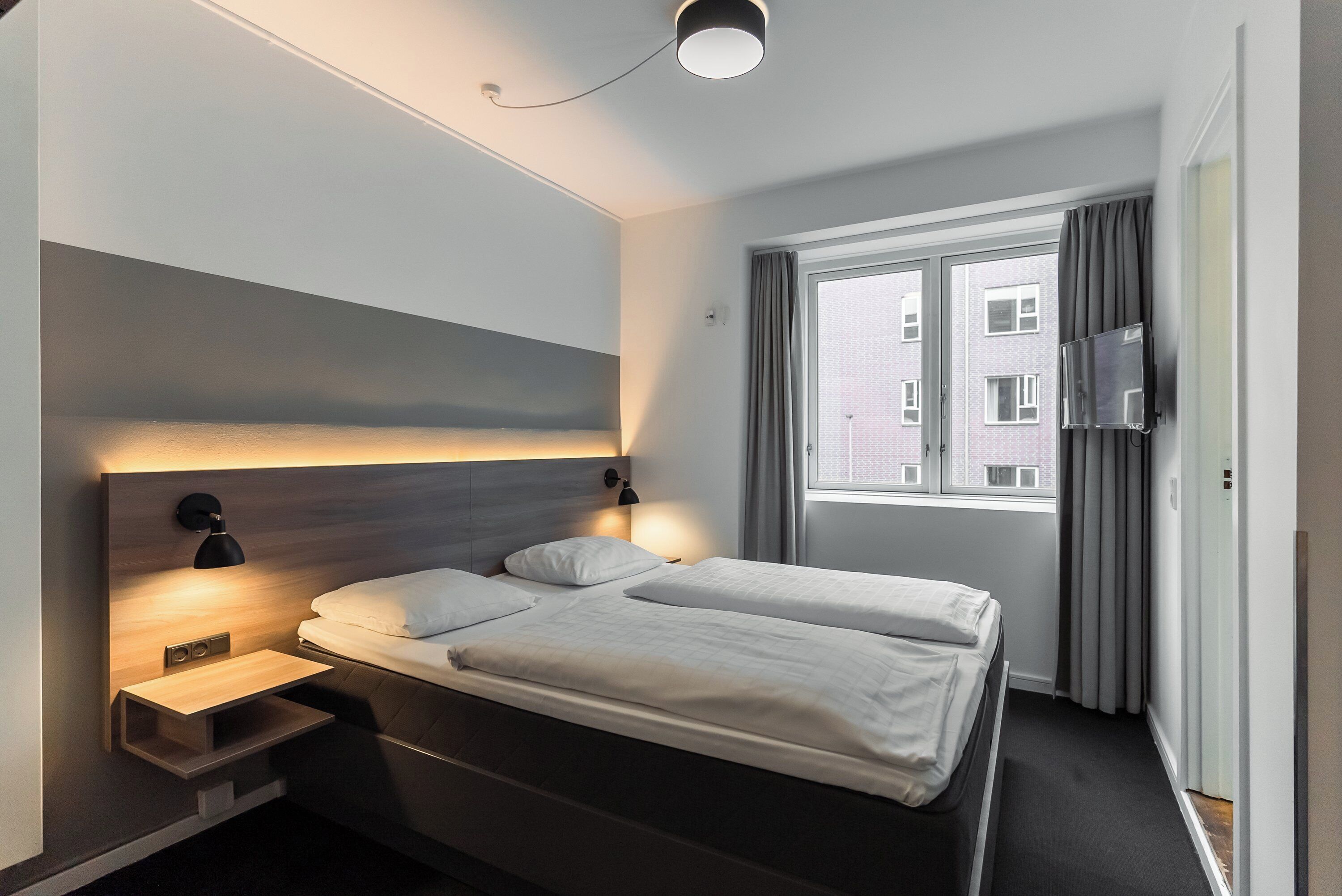 A modern hotel room with a large bed, bedside tables, a flat-screen TV, and a window with curtains.