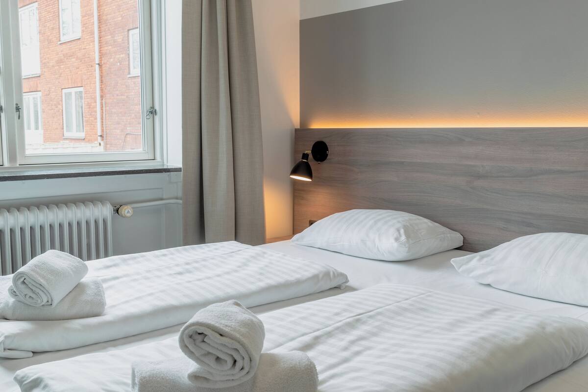 A hotel room with two beds, a window with curtains, a radiator, and a wall-mounted lamp.