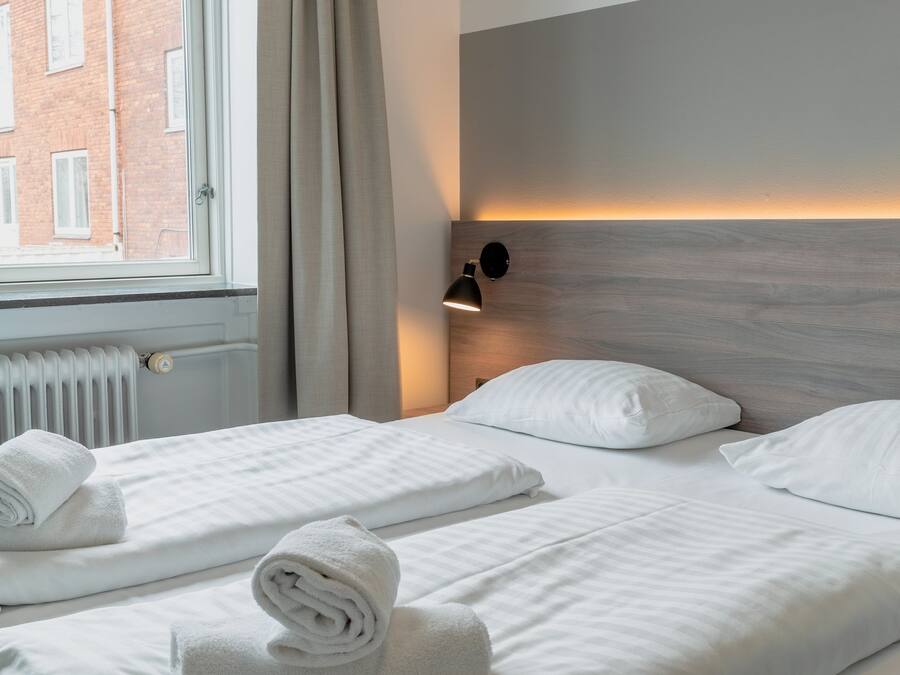 Et hotelværelse med to senge, et vindue med gardiner, en radiator og en lampe på væggen.