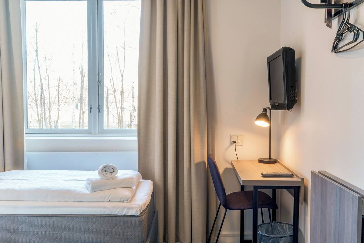 A hotel room with a bed, a desk with a lamp, a TV, and a window with curtains.