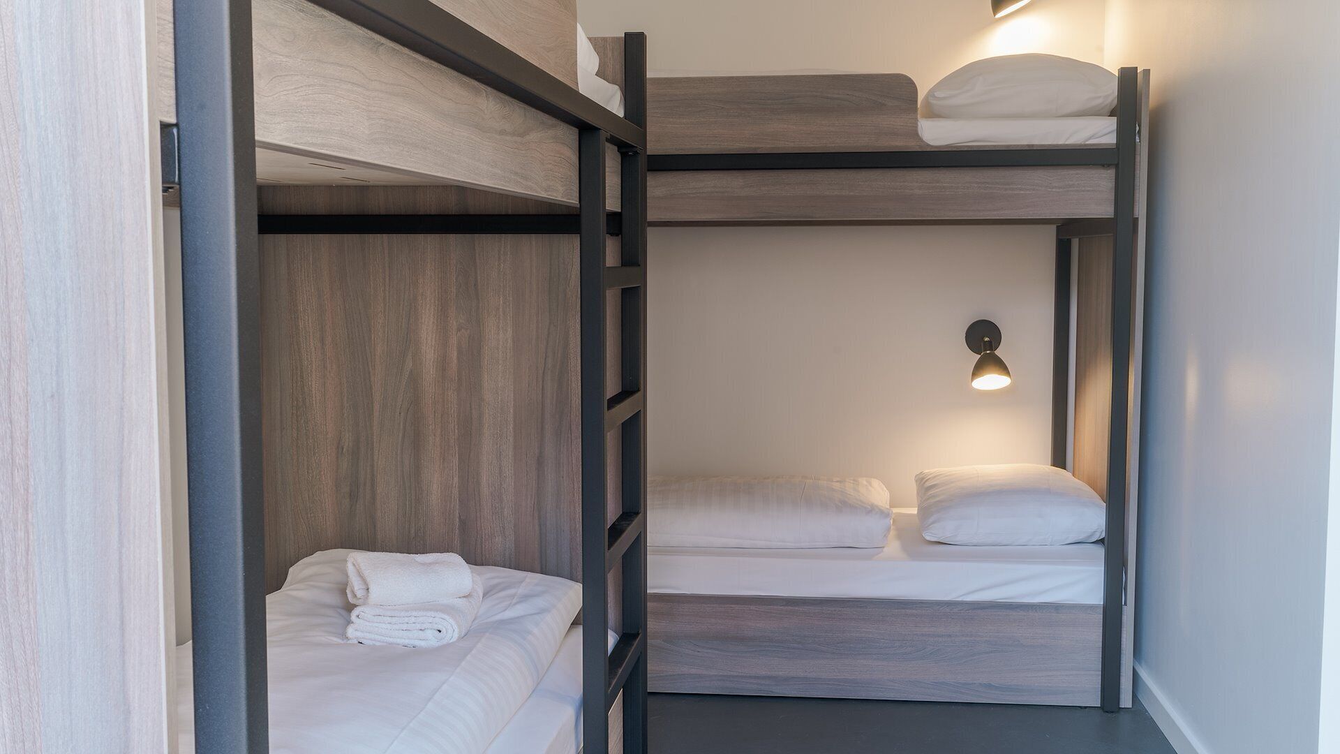 Une chambre avec un lit superposé, une tête de lit en bois, une lampe fixée au mur et une serviette soigneusement pliée sur le lit du bas.