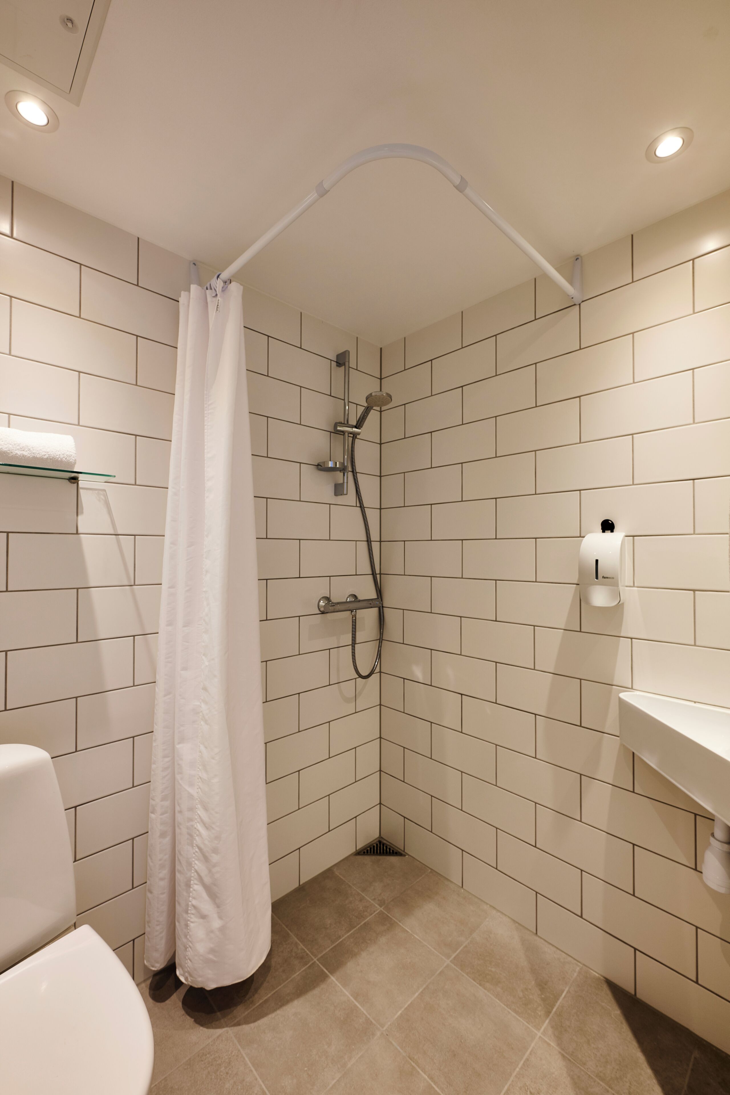 A bathroom with white tiled walls, a shower with a white curtain, a toilet, and a sink with a soap dispenser.