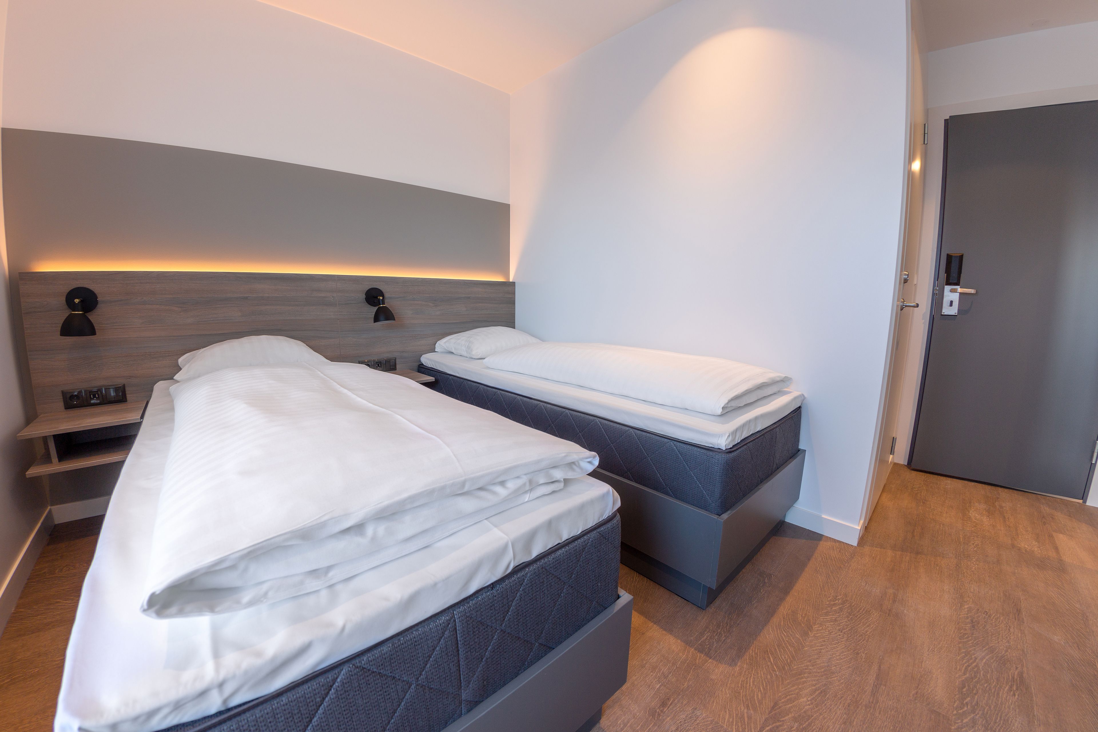 A modern hotel room with two beds, a wooden headboard, bedside lamps, and a door.