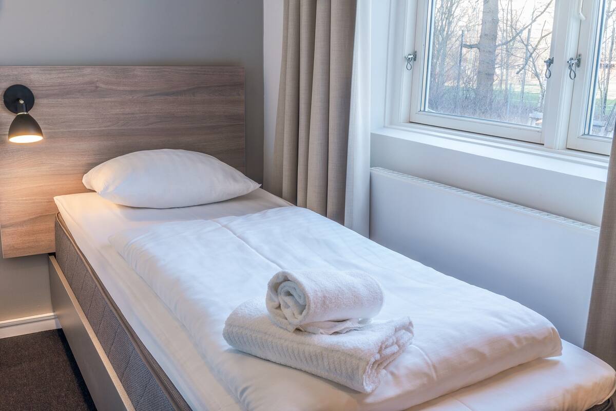 A neatly made bed with white linens and a rolled towel, a wooden headboard, a bedside lamp, and a window with curtains.