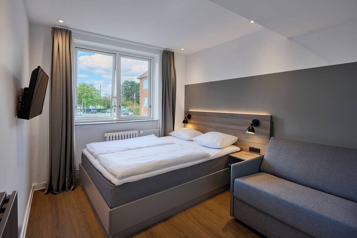 A modern hotel room with a bed, a sofa, a TV, and a window with curtains.