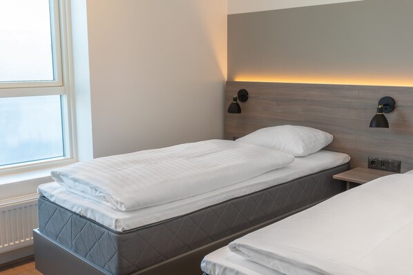 Una cama doble con colchón acolchado y ropa de cama blanca, una mesita de noche con lámpara, y una ventana con vistas al cielo.