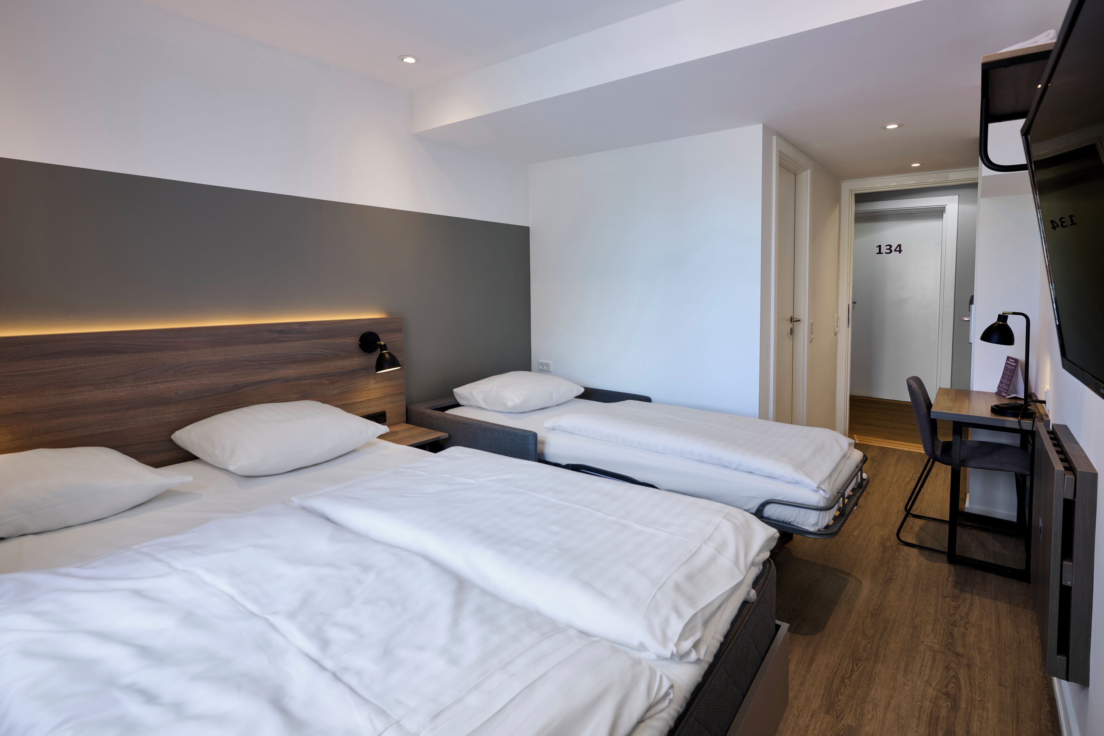 Ein Hotelzimmer mit drei Betten, einem Schreibtisch, einem Stuhl und einem Fernseher.