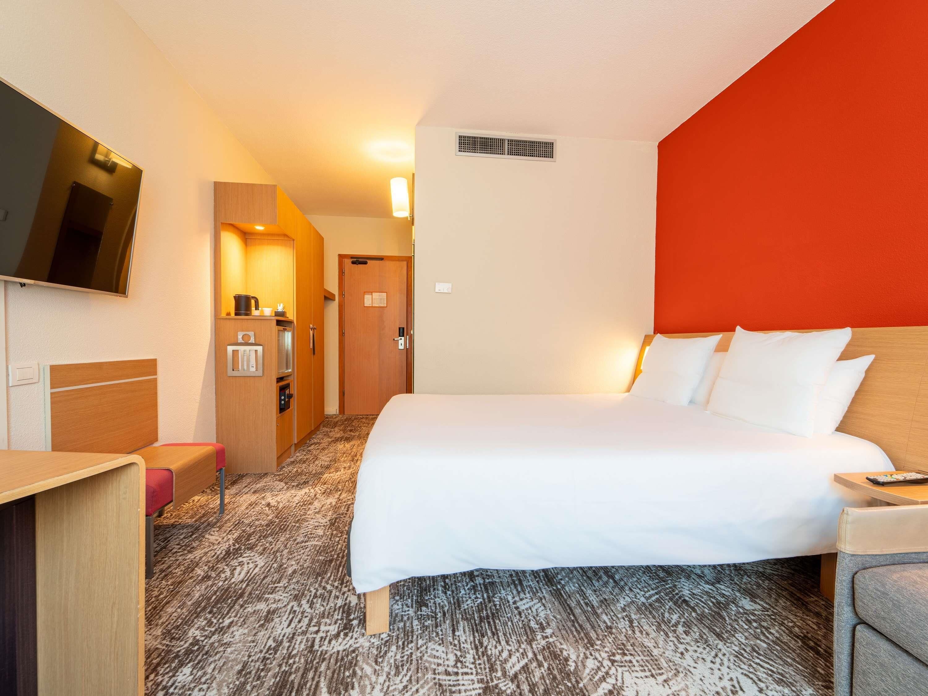 A hotel room with a bed, a desk, a television, a kitchenette, and a red accent wall.