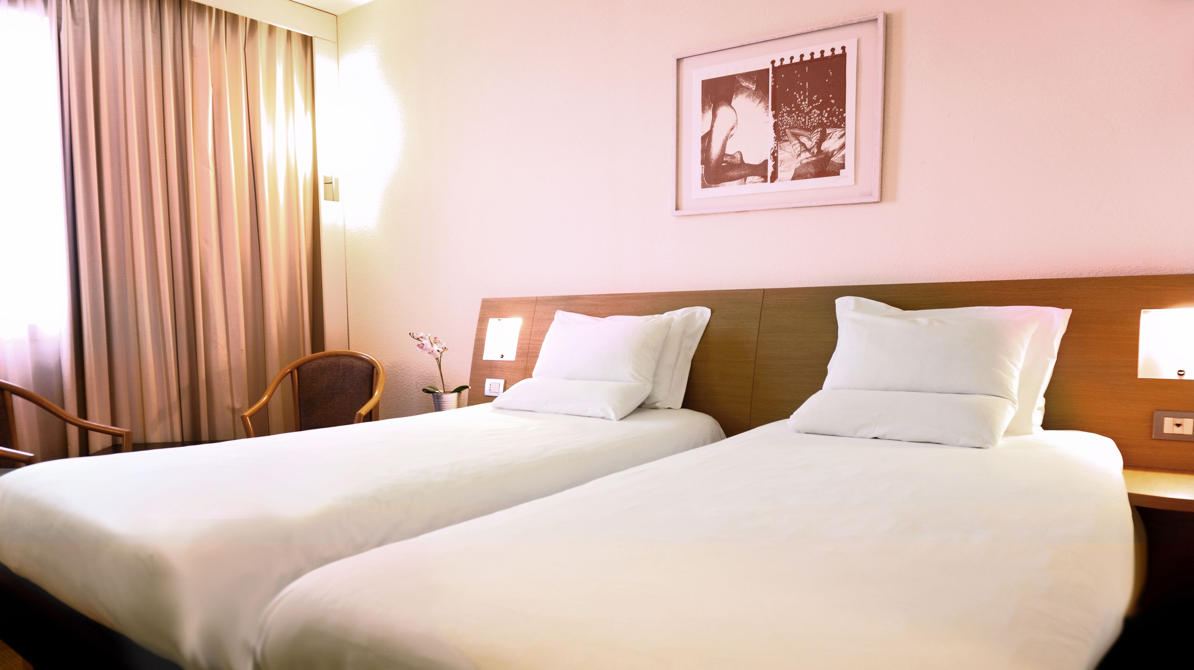 Una camera d'albergo con due letti, una testiera in legno, una sedia, un vaso con fiori e un quadro appeso al muro.