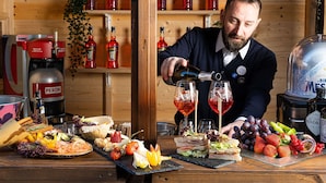 Un bar con un bartender sirviendo una bebida, comida y bebidas variadas en la barra, y estantes con botellas al fondo.