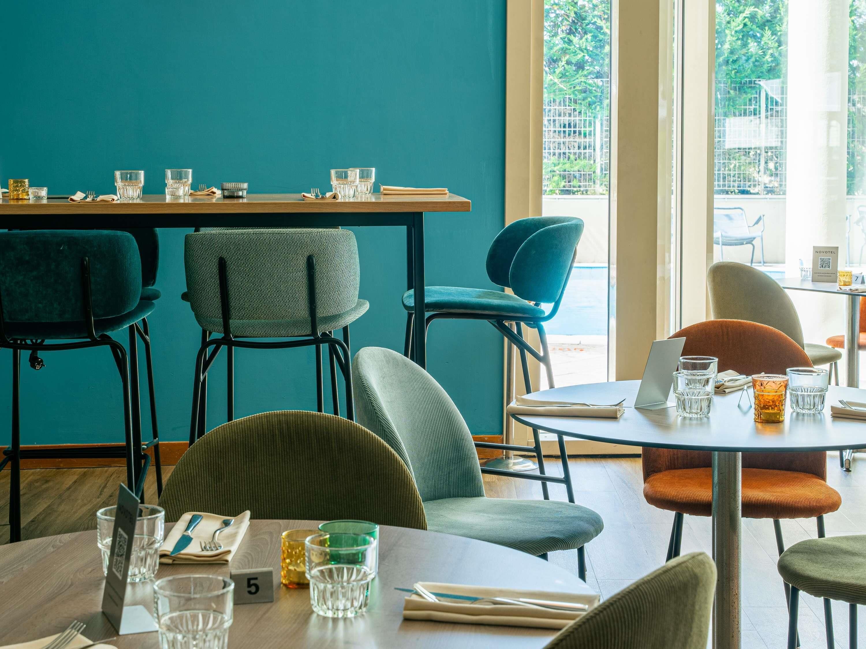 A modern dining area with teal walls, multiple seating options including chairs and a table, and a view of the outdoors through large windows.
