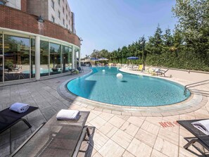 A pool area with lounge chairs, a glass-enclosed building, and a brick wall. - Novotel Roma Est (Rome)