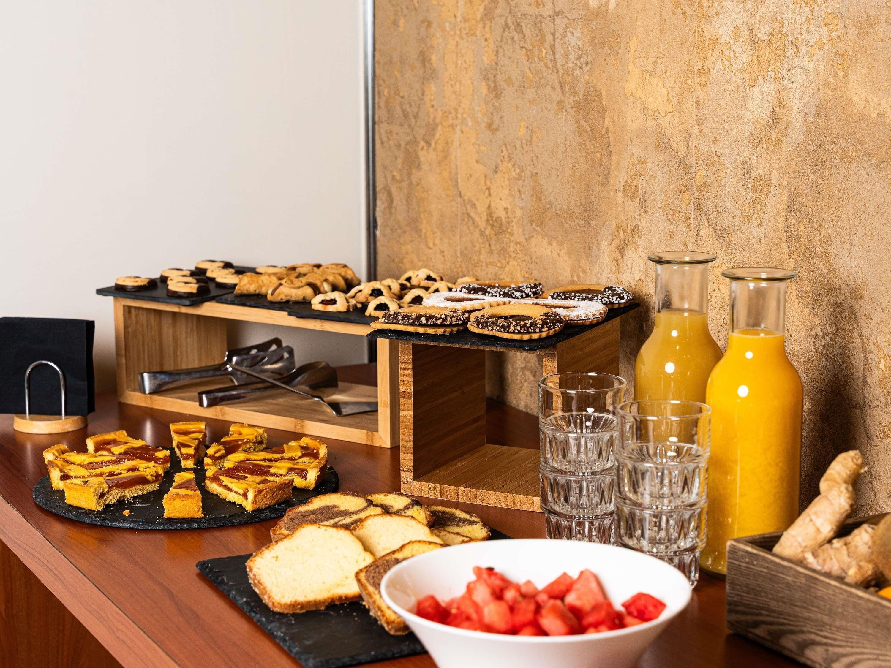 A buffet with various pastries, a slice of cake, glasses, and a bottle of orange juice.