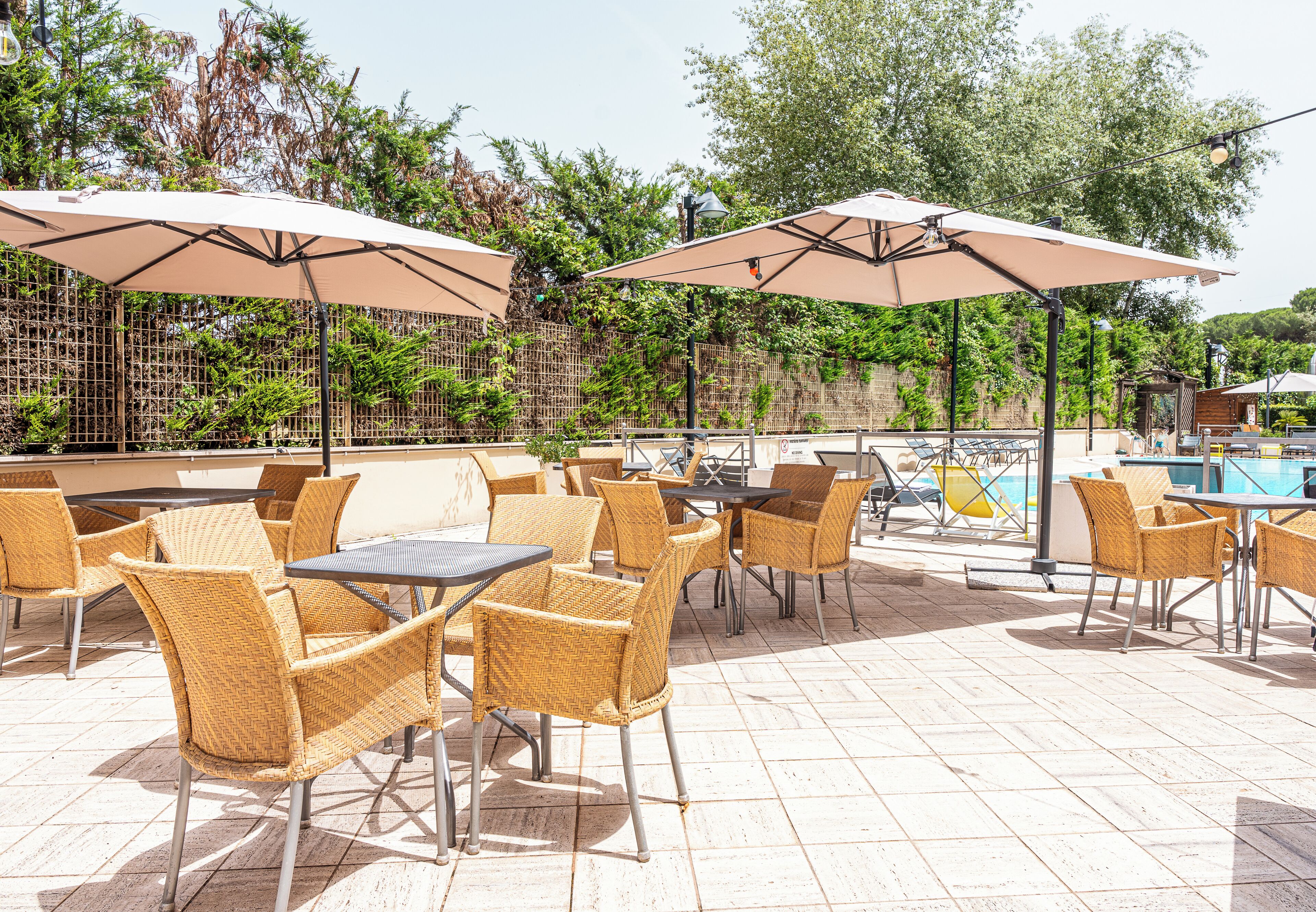 An outdoor patio area with wicker chairs and tables, umbrellas, and a pool in the background.