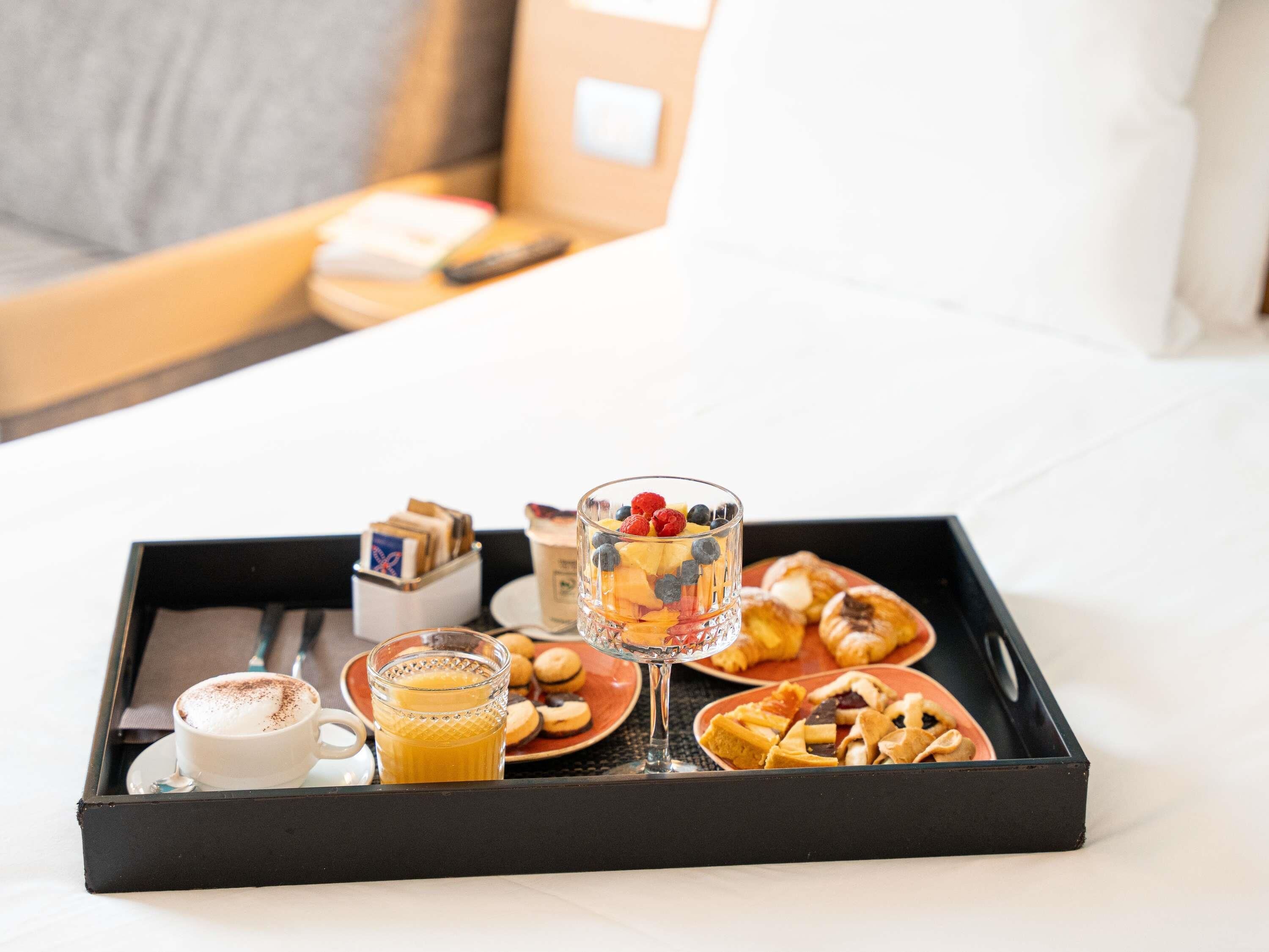 A breakfast tray with a cup of coffee, a glass of orange juice, pastries, and a fruit bowl on a bed.