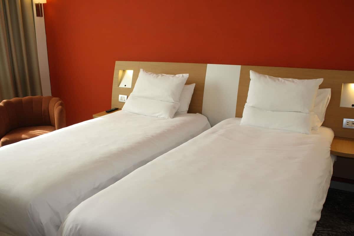 A hotel room with two single beds, a wooden headboard, a brown leather chair, and a nightstand with a lamp.