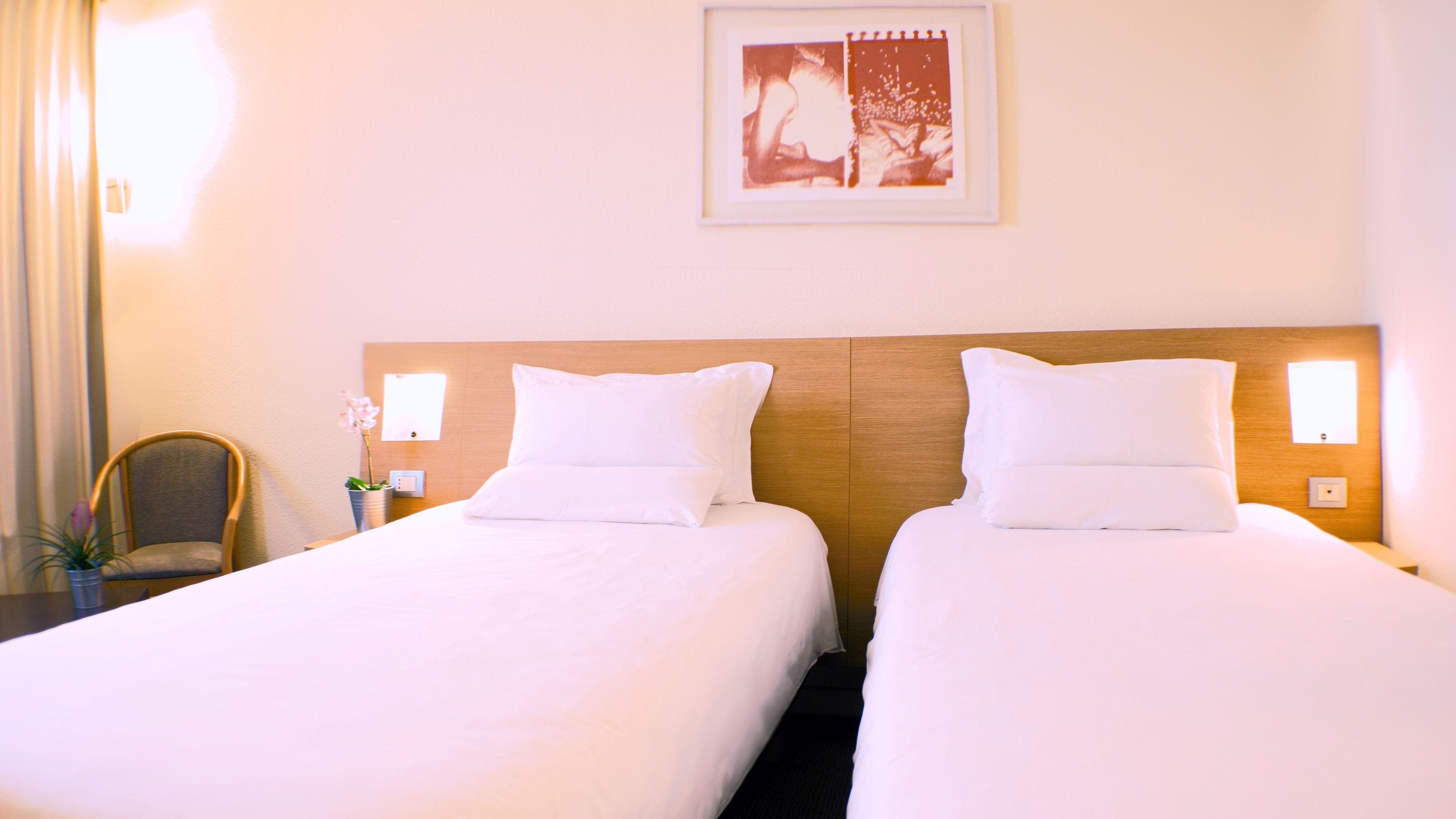 Hotellihuone, jossa on kaksi erillistä sänkyä, puiset päätyosat, valkoiset vuodevaatteet, pieni pöytä, jossa on maljakko täynnä kukkia, ja seinällä oleva kehystetty kuva.