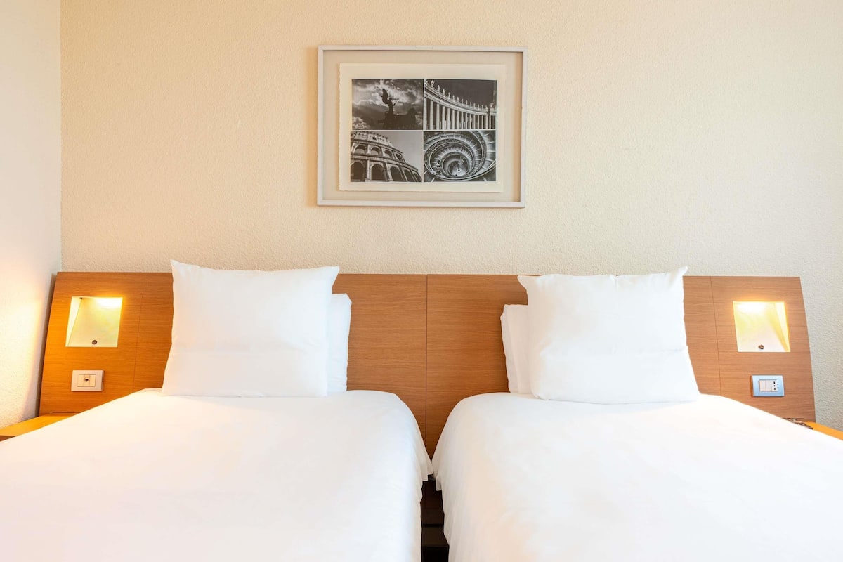A hotel room with two beds, each with a wooden headboard and bedside lamps. A framed picture hangs on the wall above the beds.