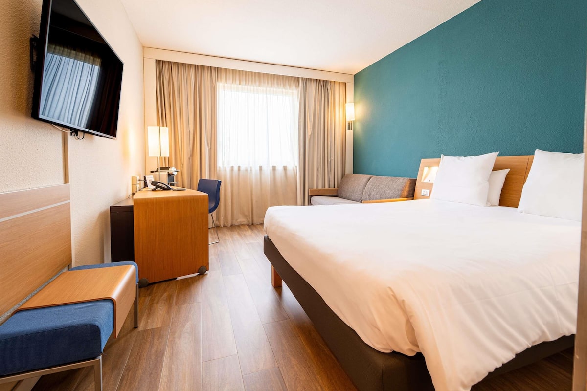 A hotel room with a bed, a desk, a chair, a TV, and a window with curtains.