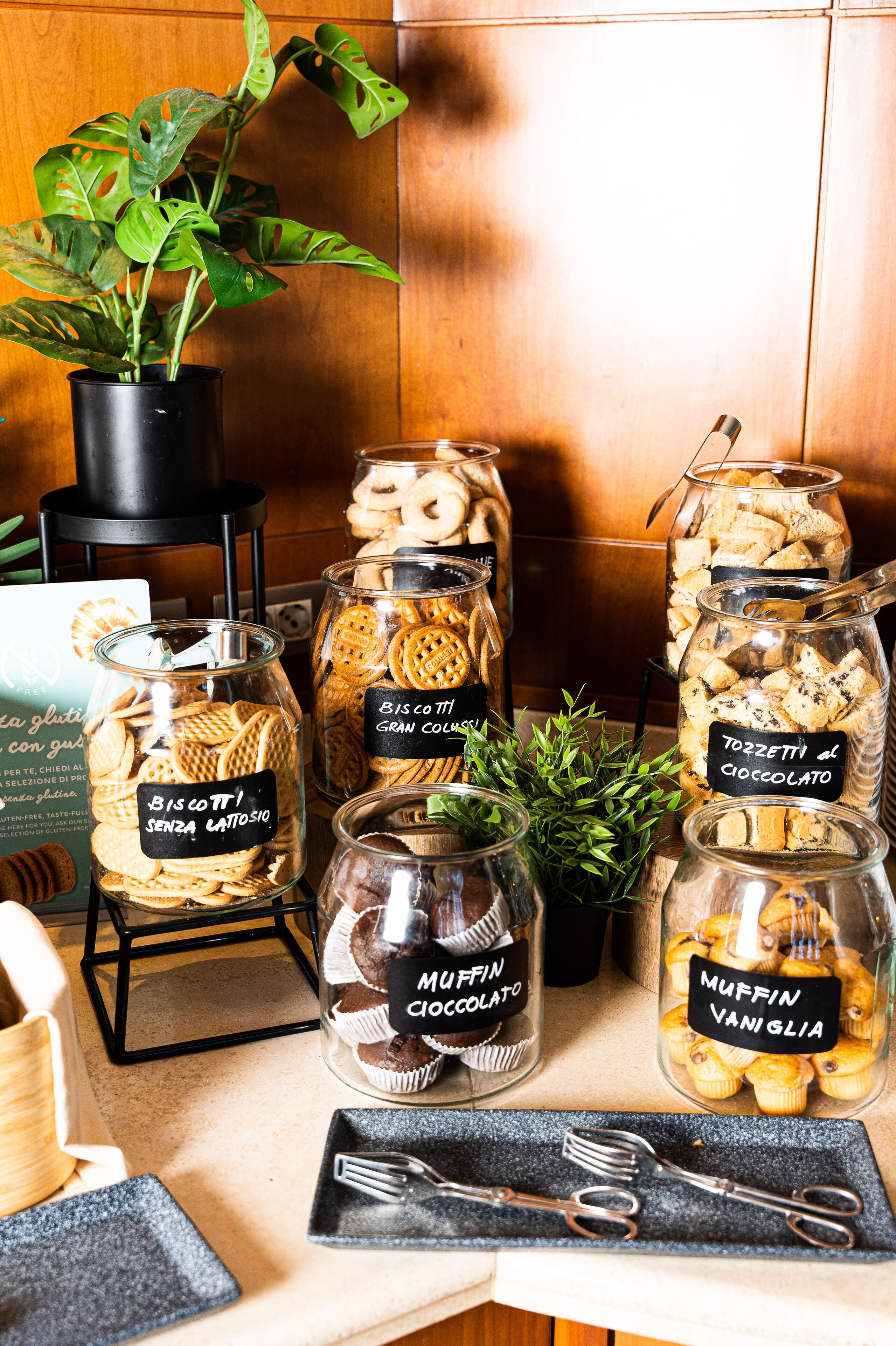 Exposição de diversos produtos de panificação em potes de vidro, incluindo biscoitos e muffins, com etiquetas que indicam sabores como chocolate e baunilha.
