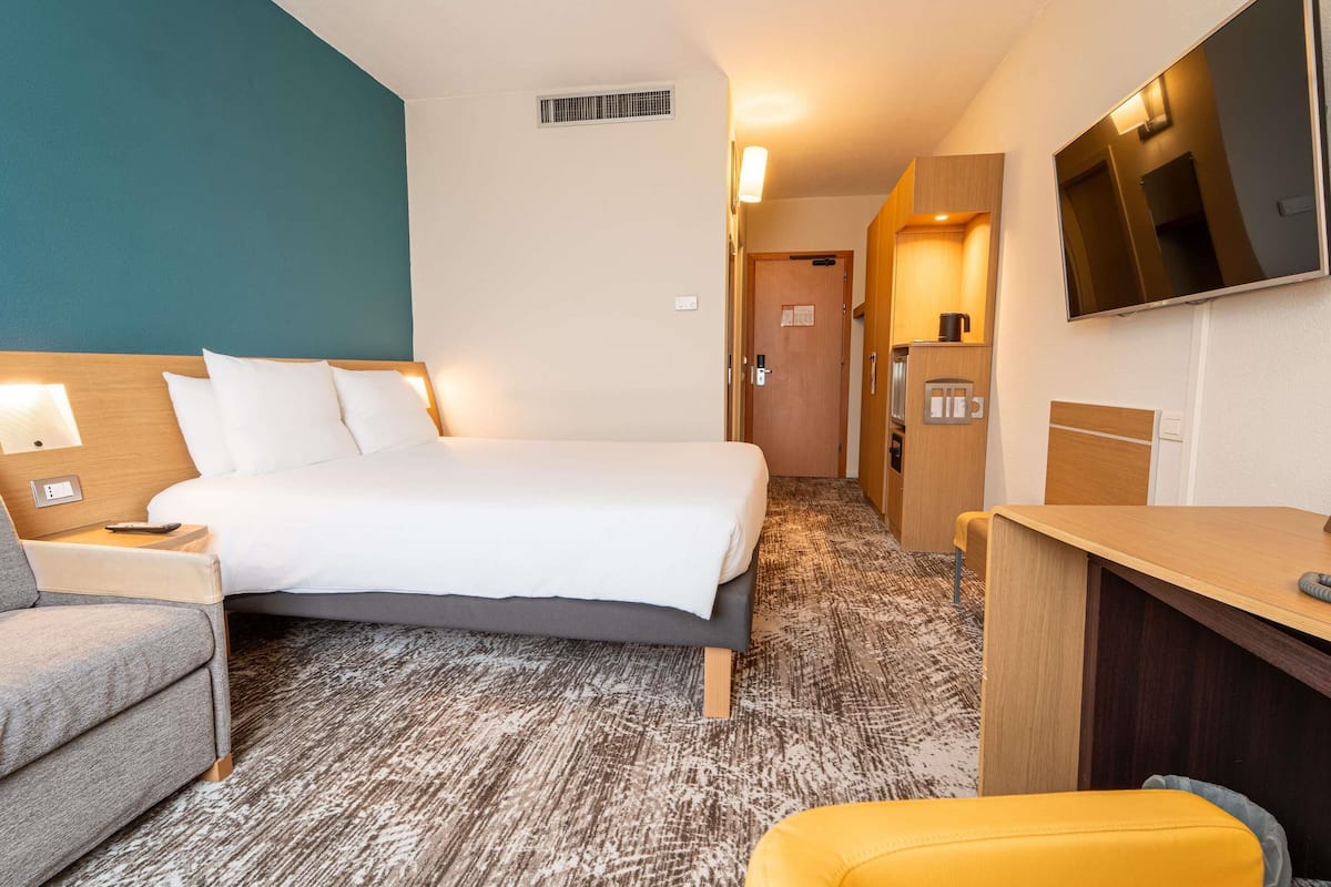 A hotel room with a bed, a desk, a chair, a TV, and a kitchenette.