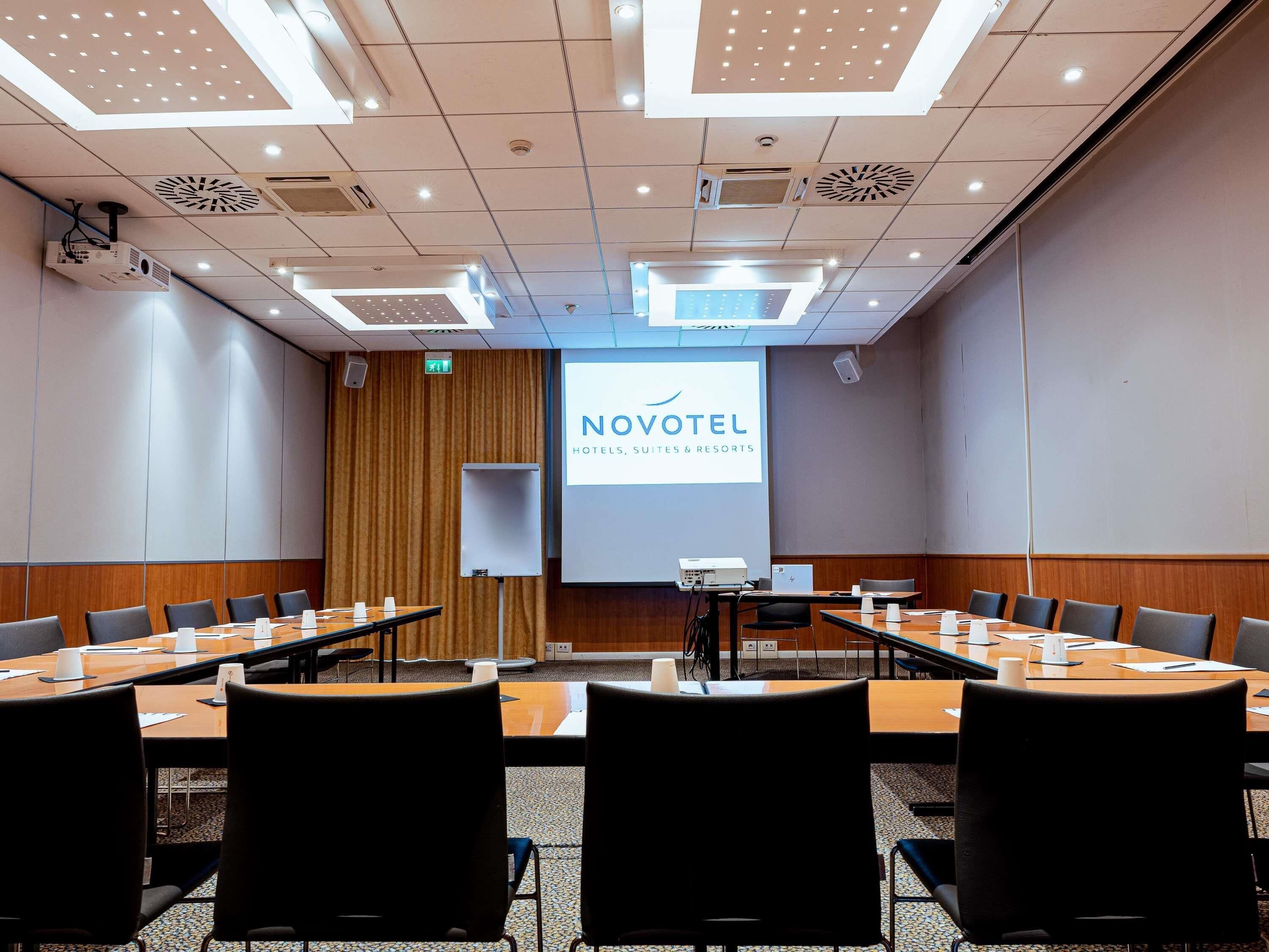 Un salón de conferencias con una pantalla de proyector mostrando el logotipo de NOVOTEL HOTELS, SUITES & RESORTS, varias mesas con sillas y una pared de acento de madera.