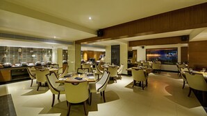 Um restaurante espaçoso com várias mesas e cadeiras, uma área de cozinha e uma tela grande exibindo uma paisagem.