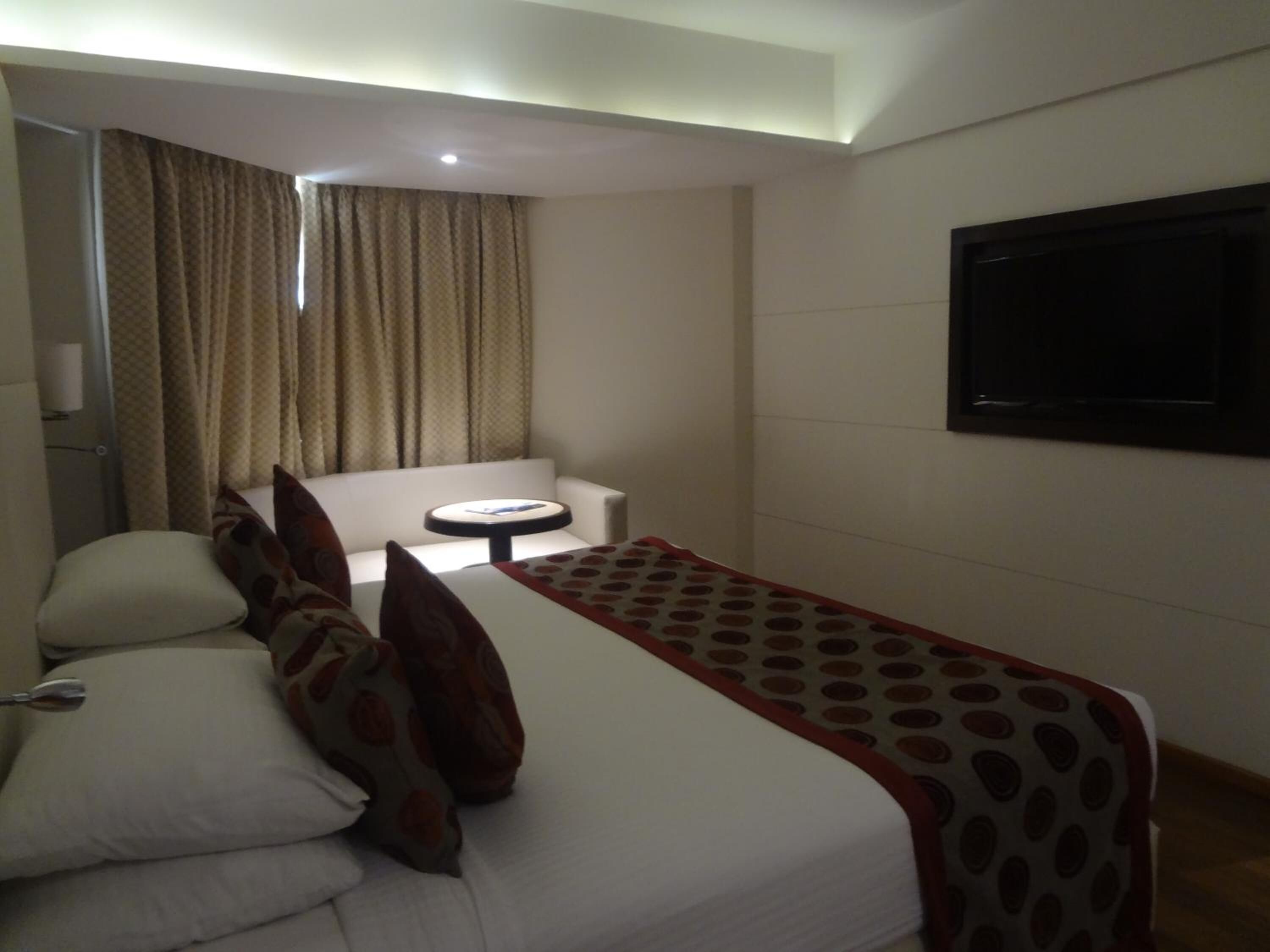 Una habitación de hotel con cama, una mesita, una silla, televisor y cortinas.