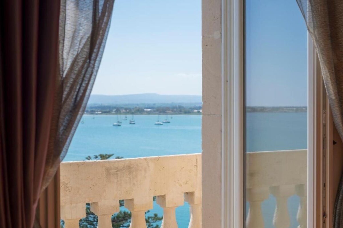 A view through a window with curtains, a balcony, and a body of water with boats.