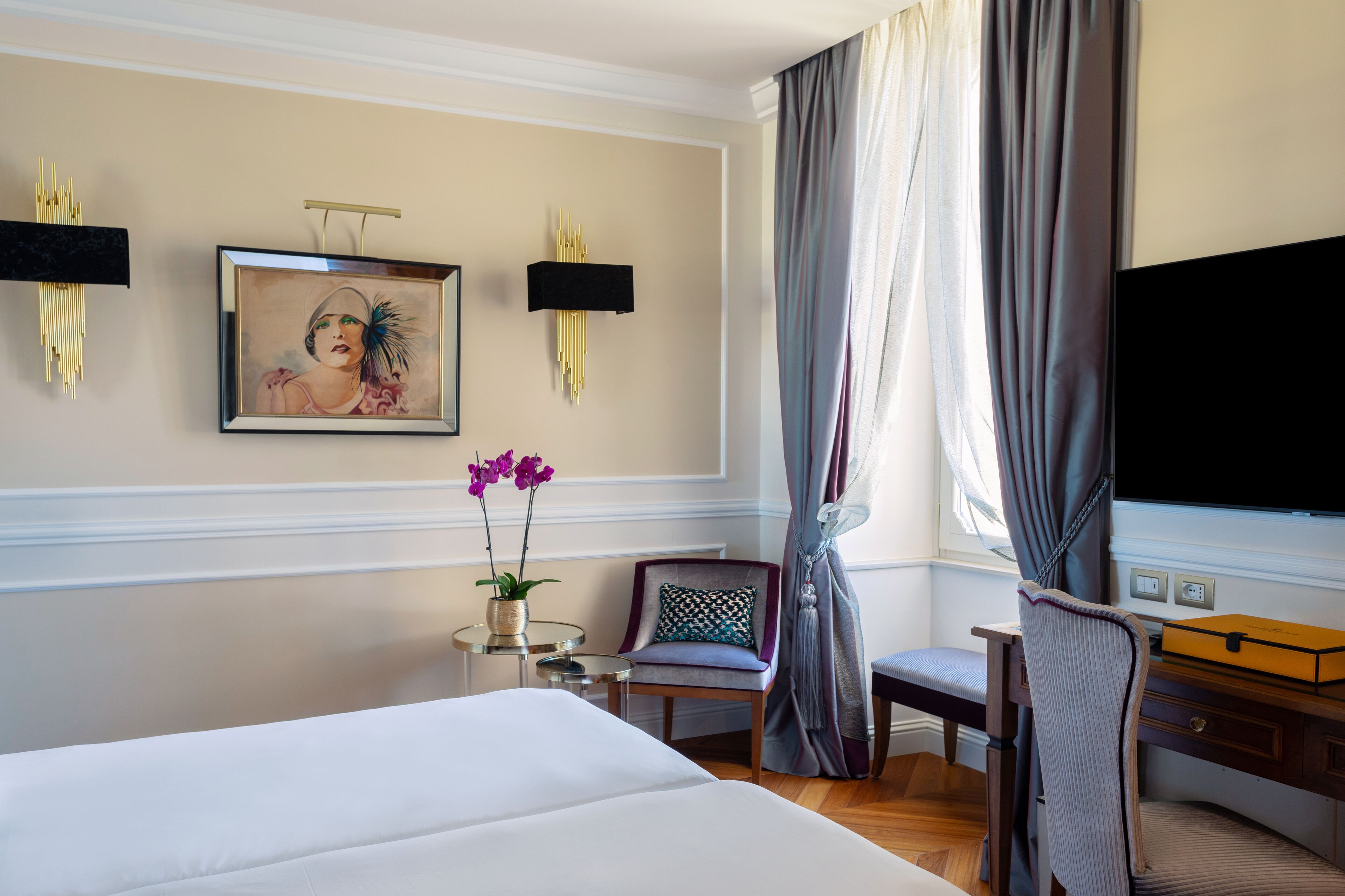 Ein Hotelzimmer mit Bett, Schreibtisch, Sessel, Fernseher und einem Gemälde an der Wand.