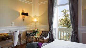 A hotel room with a bed, a desk, two chairs, a table with flowers, a lamp, and a view of trees through a window.