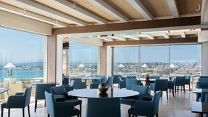 A spacious dining area with round tables and blue chairs, large windows offering a city view, and a wooden ceiling structure. - Grand Hotel Des Ètrangers - The Leading Hotels of the World (Syracuse)
