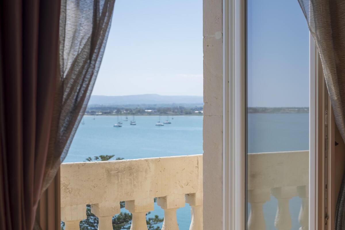 A view from a balcony overlooking a body of water with boats, a distant landscape, and a building in the background.
