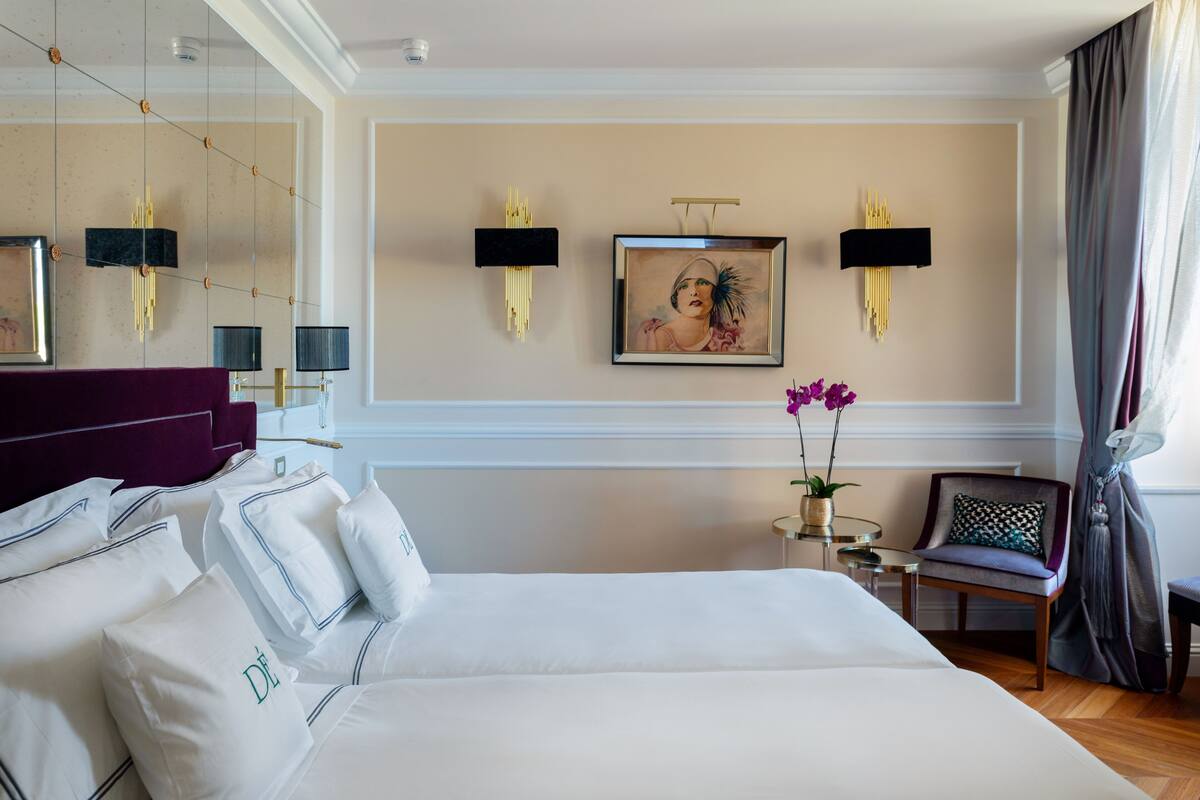 A hotel room with a large bed, a small table, a chair, and wall-mounted lamps.