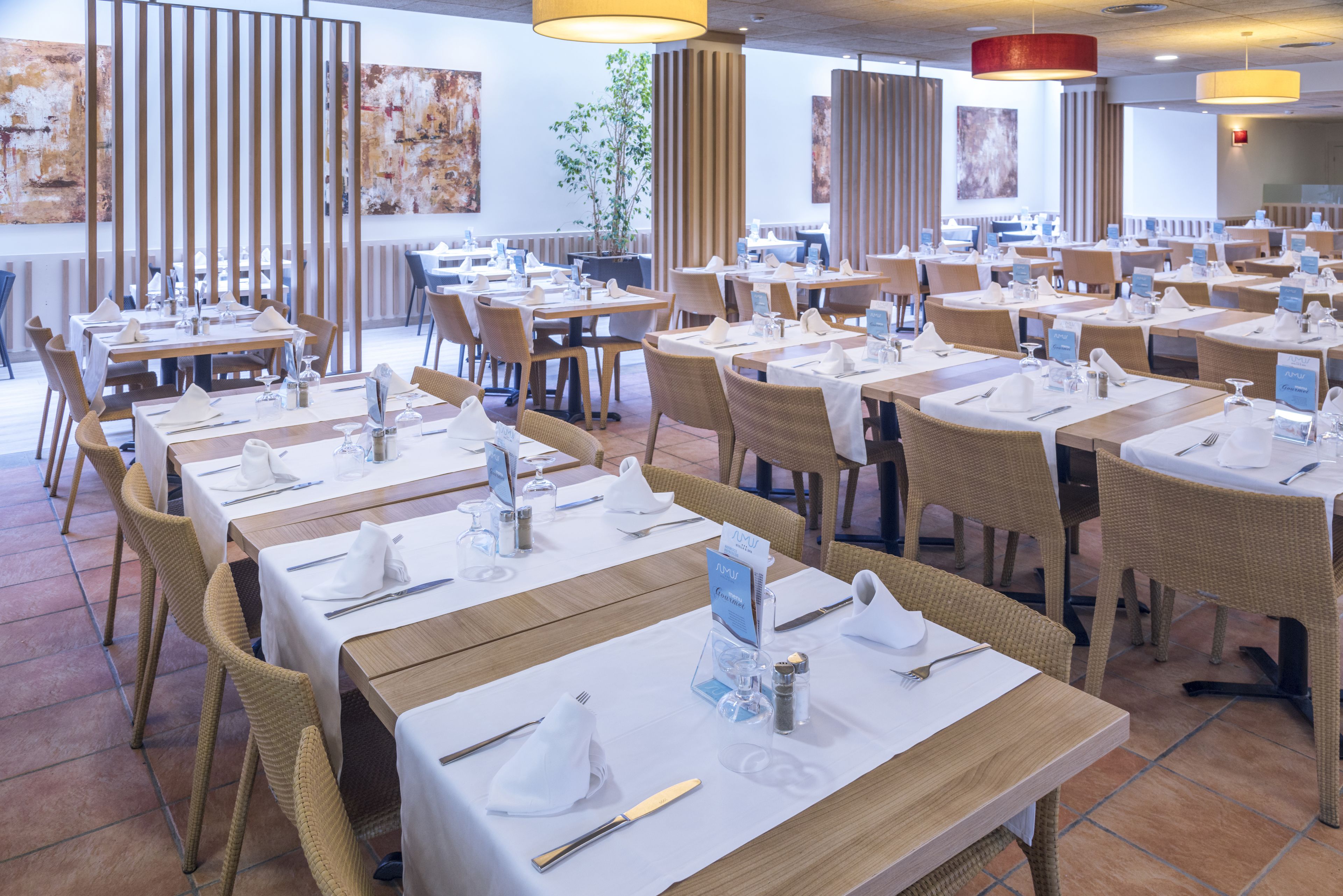 Un restaurante con varias mesas dispuestas para comer, con manteles blancos, sillas de mimbre e iluminación colgante moderna.
