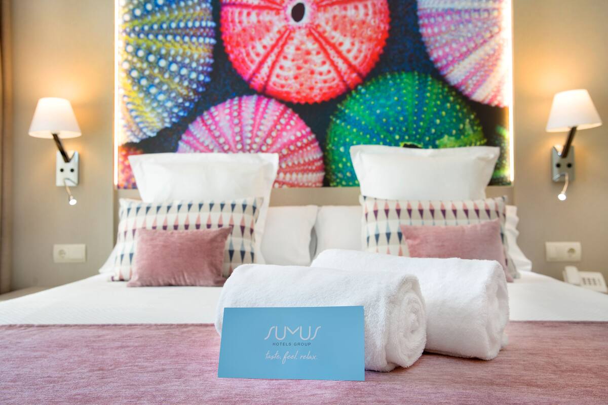 A hotel room with a bed, pillows, a rolled towel, and a wall painting.