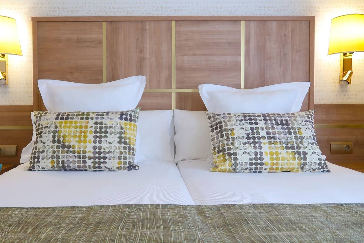 A double bed with two pillows, a wooden headboard, and two wall-mounted lamps.