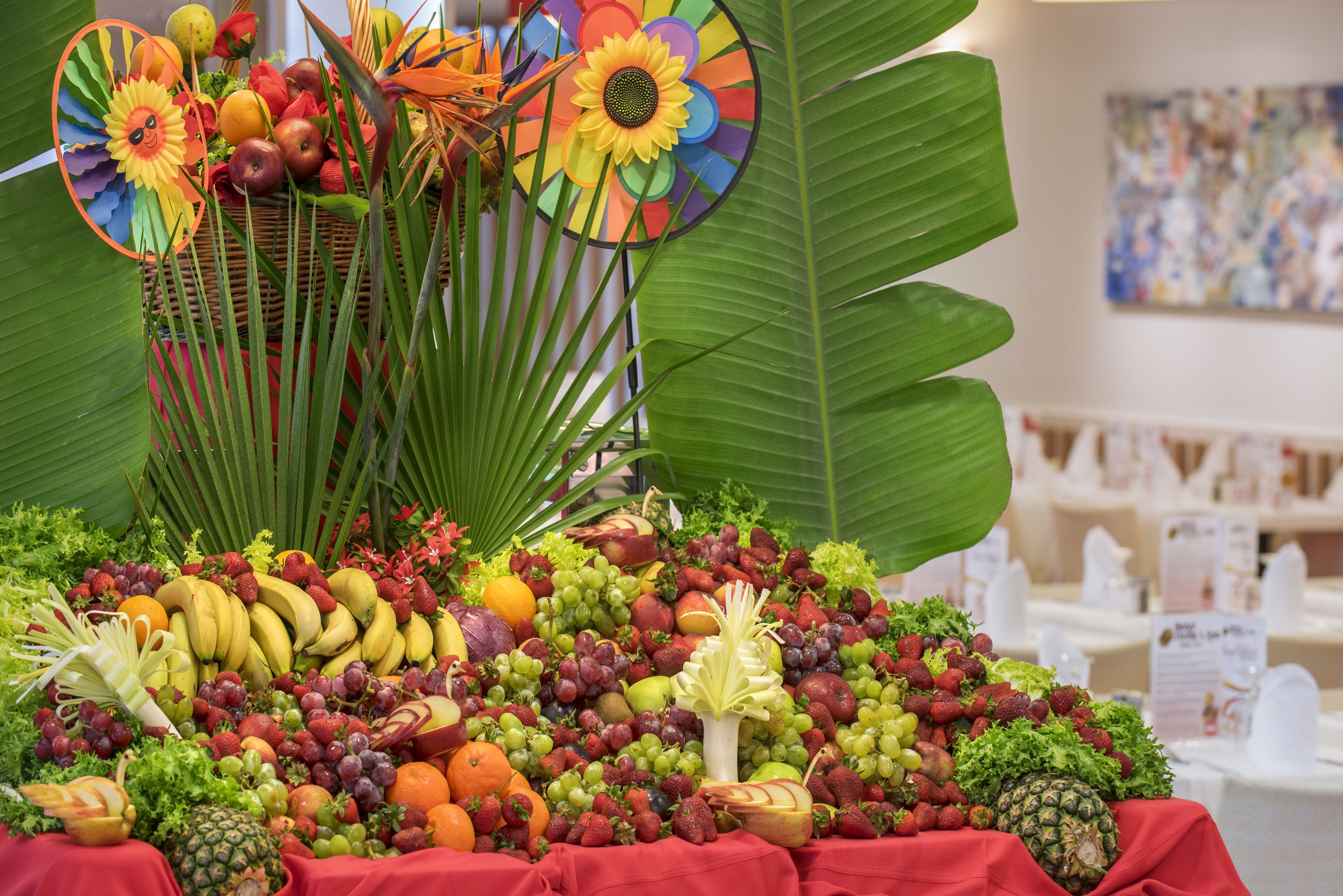 Exhibición de frutas y verduras con mantel rojo, rodeada de hojas verdes grandes y flores de papel decorativas.