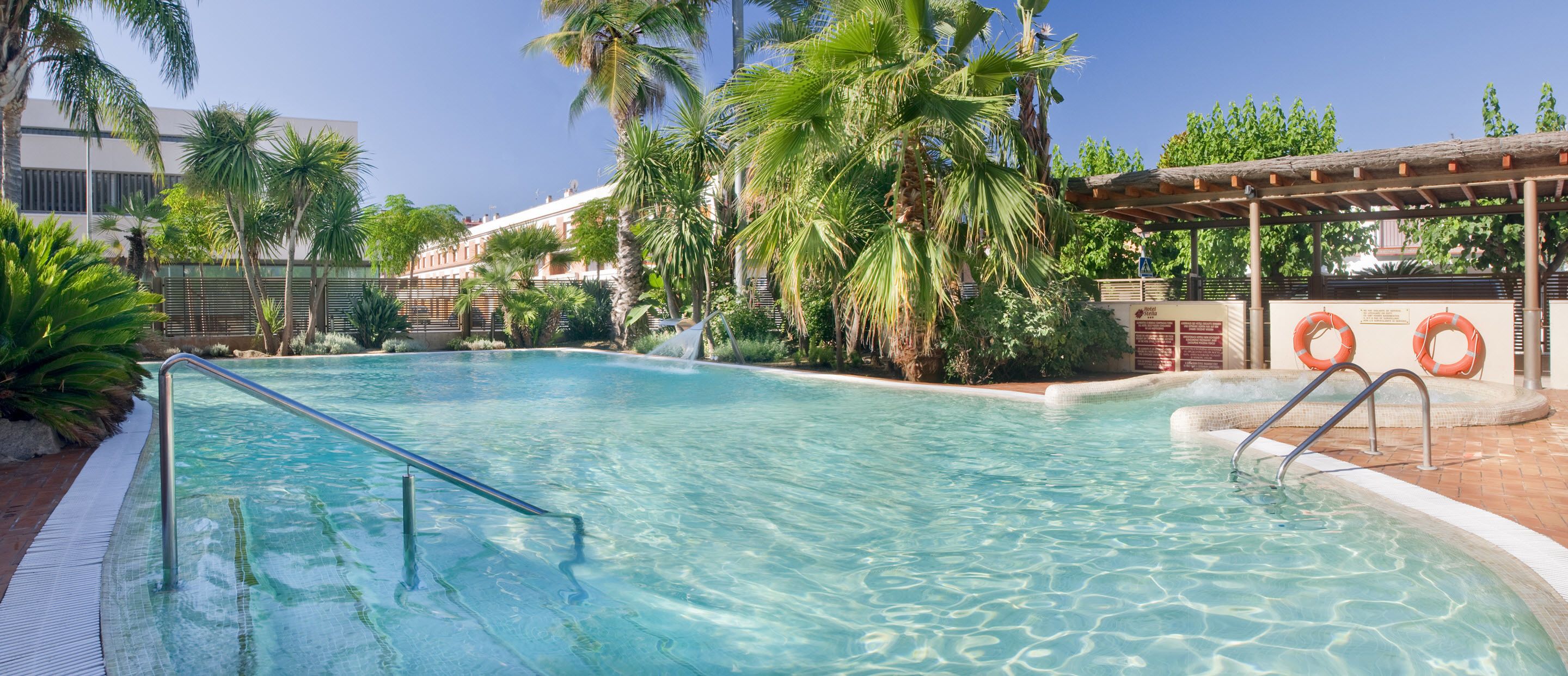 A clear swimming pool with a metal ladder, surrounded by palm trees and a pergola.