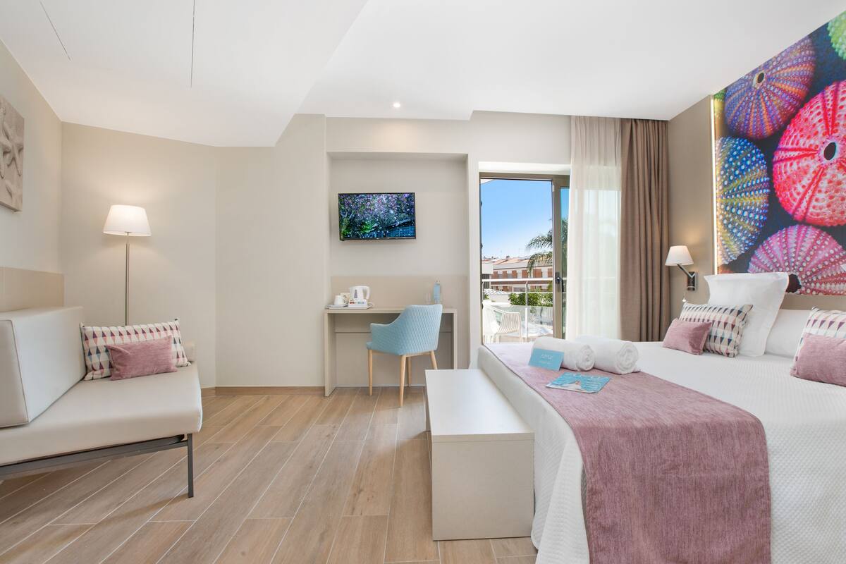 A hotel room with a bed, a desk, a chair, a sofa, and a large window with a view.