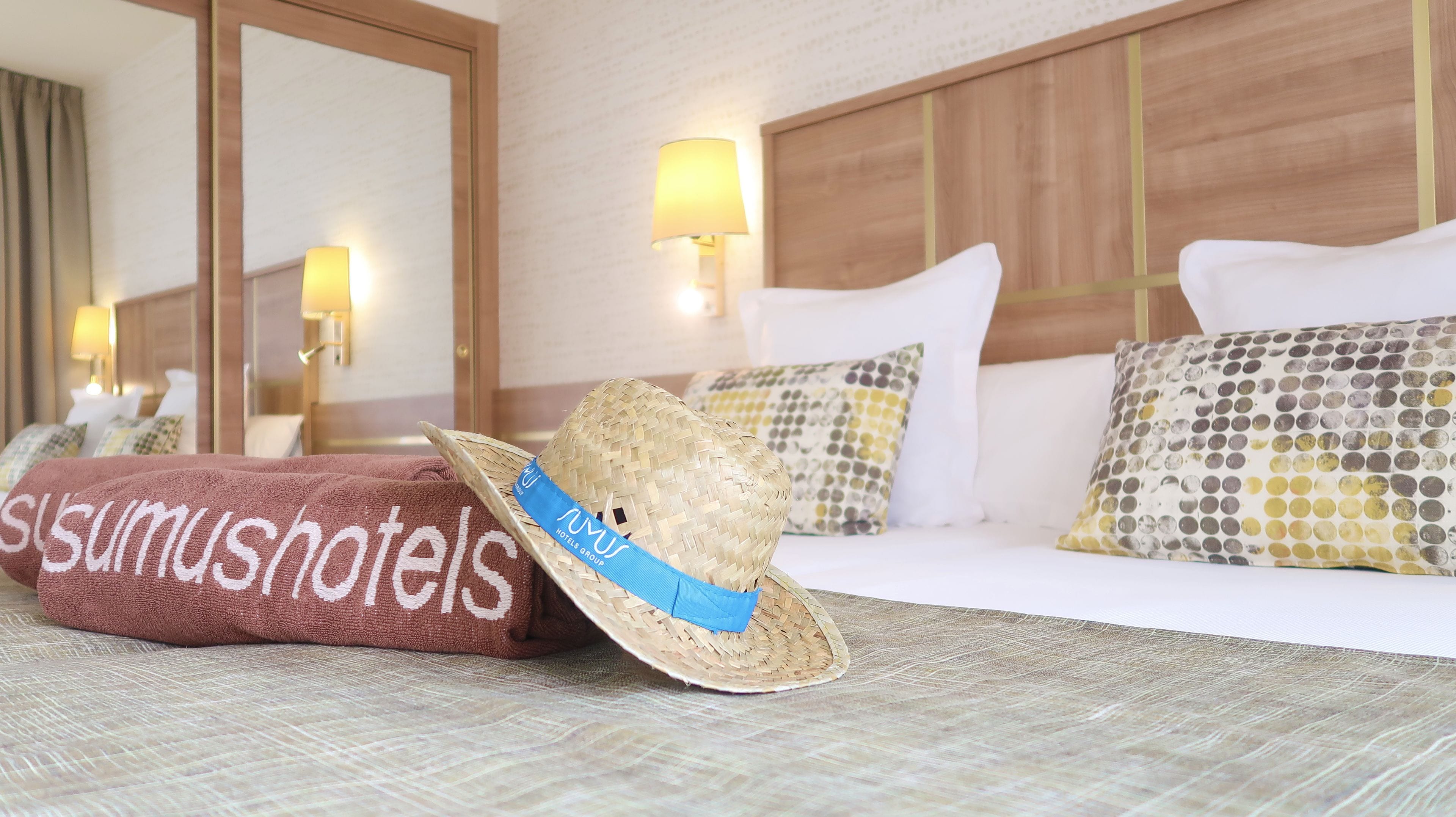 Habitación de hotel con cama, sombrero tejido y toalla con el nombre del hotel.