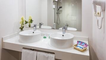 Un baño con dos lavamanos, un espejo, un secador de pelo y una cesta de artículos de aseo.
